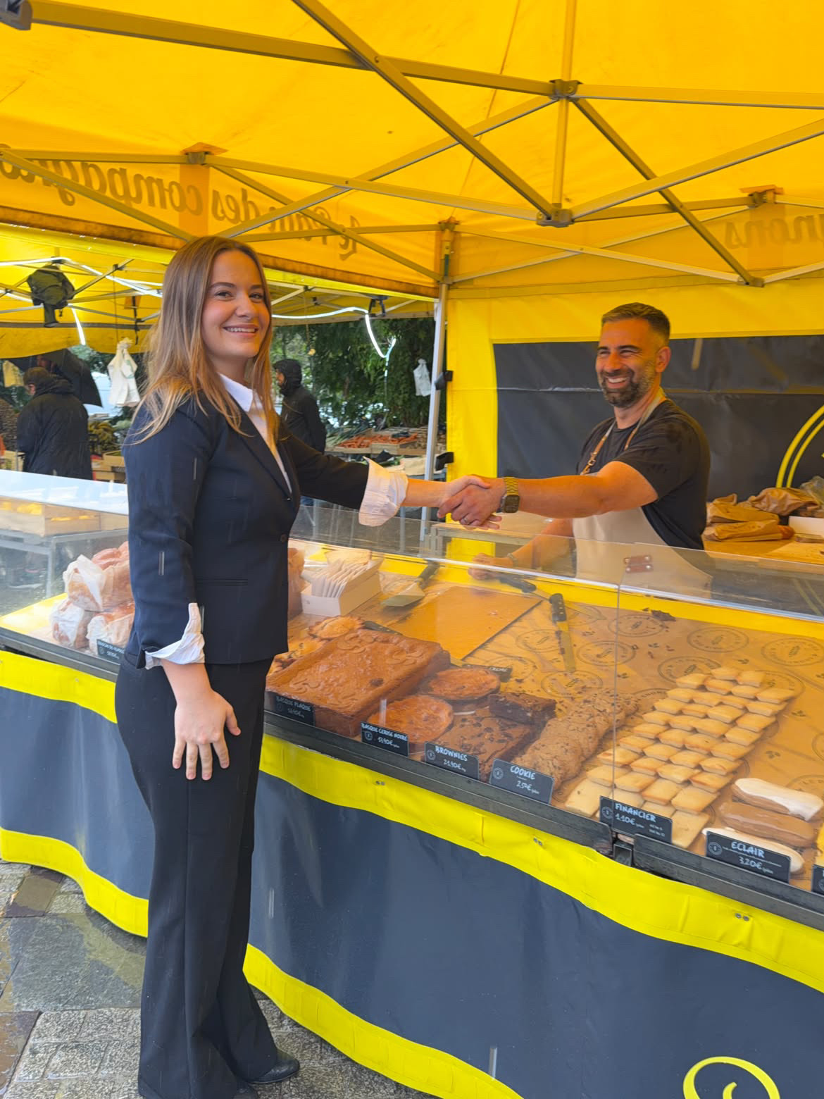 Clara Caurier échange une poignée de main avec un commerçant devant un étal de pâtisseries